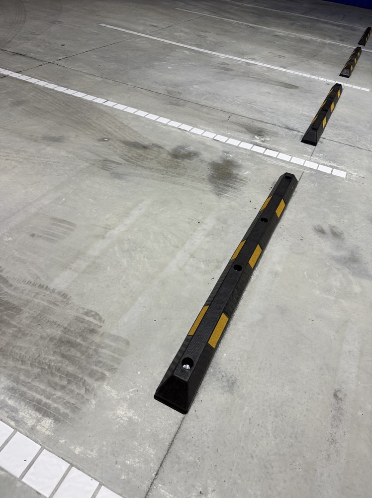 Butée de parking : sécurité et délimitation sur sol béton. Butées de parking noires avec bandes réfléchissantes jaunes installées sur un sol en béton gris marqué par des lignes blanches de stationnement.