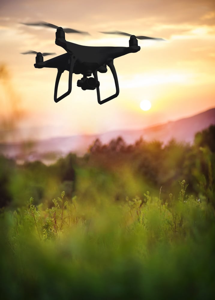 Drone en vol : Paysage nature au coucher de soleil Quadcopter noir volant au crépuscule sur un paysage de forêt et de montagnes, herbe verte floue au premier plan.