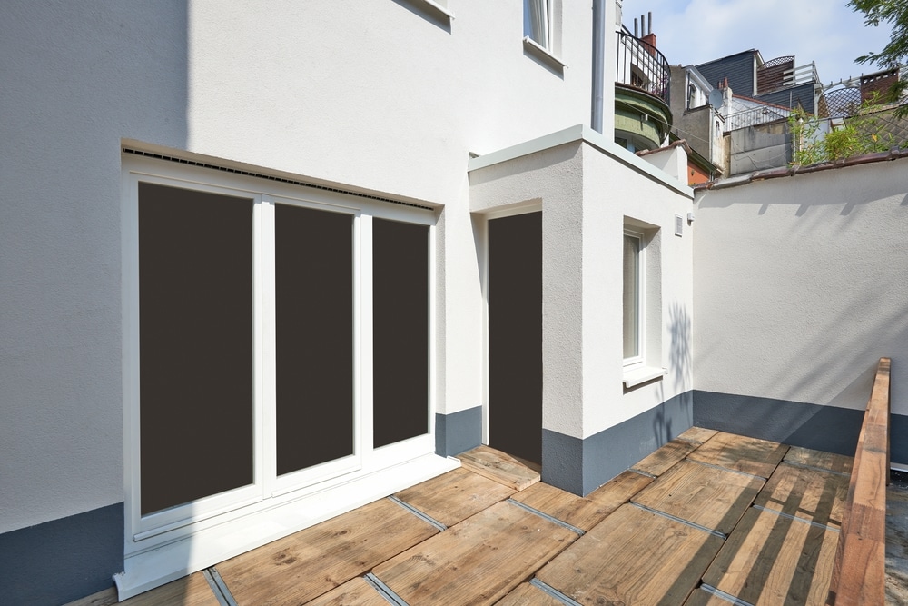 Terrasse urbaine en bois, baie vitrée et façade blanche Grande baie vitrée et façade blanche avec soubassement gris. Terrasse en bois dans une cour arrière urbaine, sous un ciel bleu.