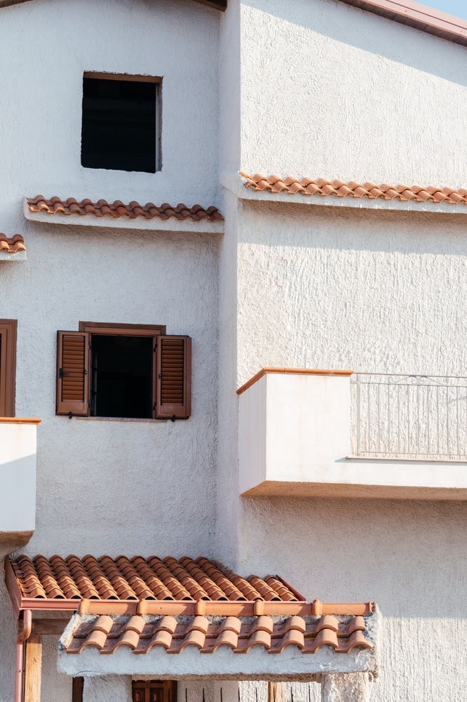 Façade méditerranéenne: crépi blanc, tuiles et volets bois. Façade moderne en crépi blanc avec volets bois marron ouverts, petites toitures en tuiles terracotta et balcon ensoleillé.