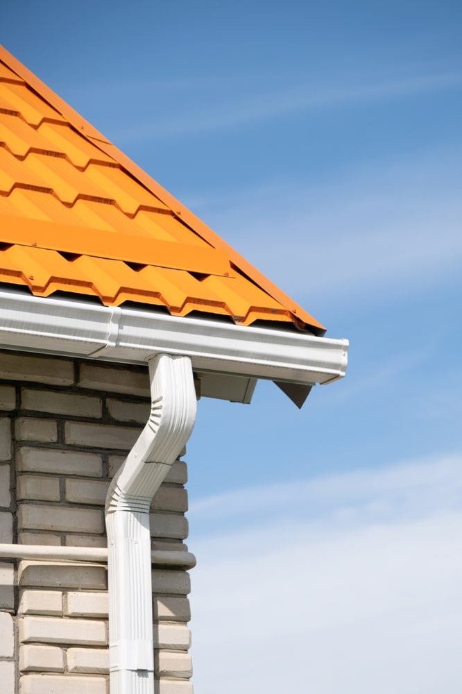 Gouttière et toiture orange : système d'évacuation des eaux Toiture orange moderne en tôle ondulée. Système de gouttière et descente d'eau blancs fixés sur le mur en briques claires.