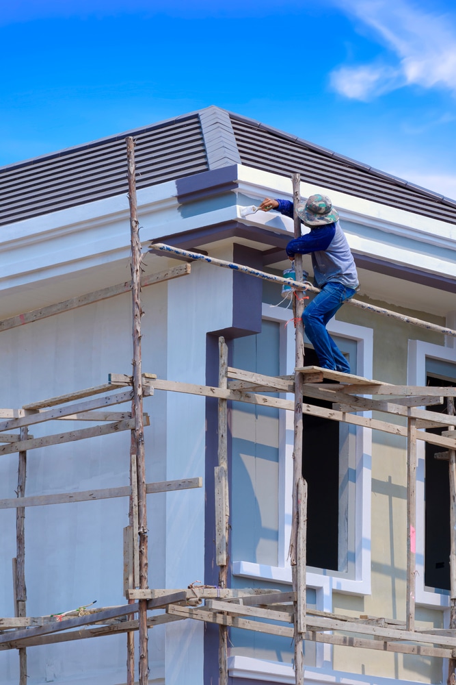 Peintre en bâtiment : Travaux de finition sur échafaudage Ouvrier sur échafaudage en bois peignant la corniche d'une maison neuve en blanc sous un ciel bleu vif. Construction.