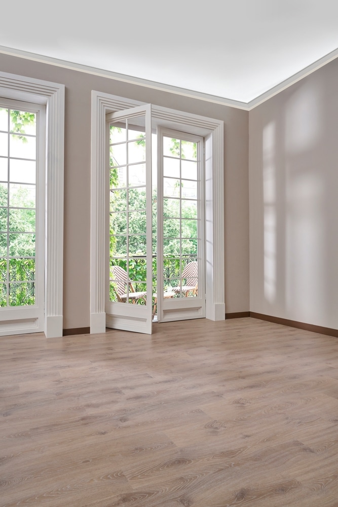 Pièce élégante : parquet stratifié clair et moulures blanches Pièce élégante aux murs taupe et parquet clair. Porte-fenêtre à grille et moulures blanches donnant sur un balcon arboré avec chaises.