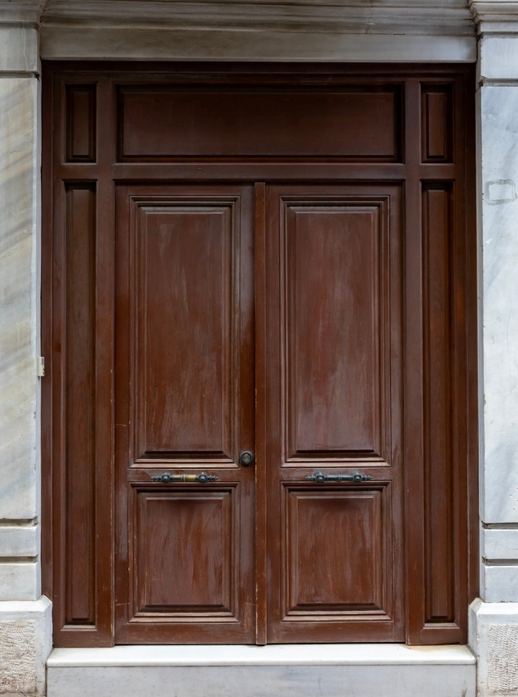 Élégante porte double en bois marron classique et marbre Porte double classique en bois marron foncé à panneaux, encadrée par des montants en marbre blanc veiné et surmontée d'un linteau mouluré.
