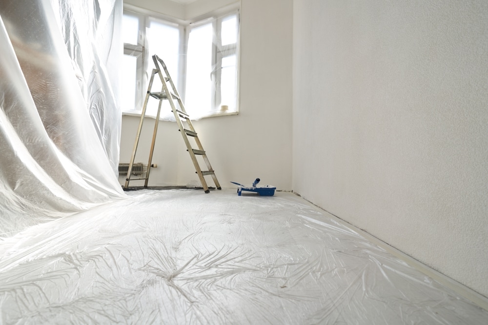 Travaux de peinture : Préparation de la pièce et protection. Rénovation d'une pièce : sol couvert de bâche plastique, échelle près de la fenêtre, murs blancs texturés et rouleau de peinture.
