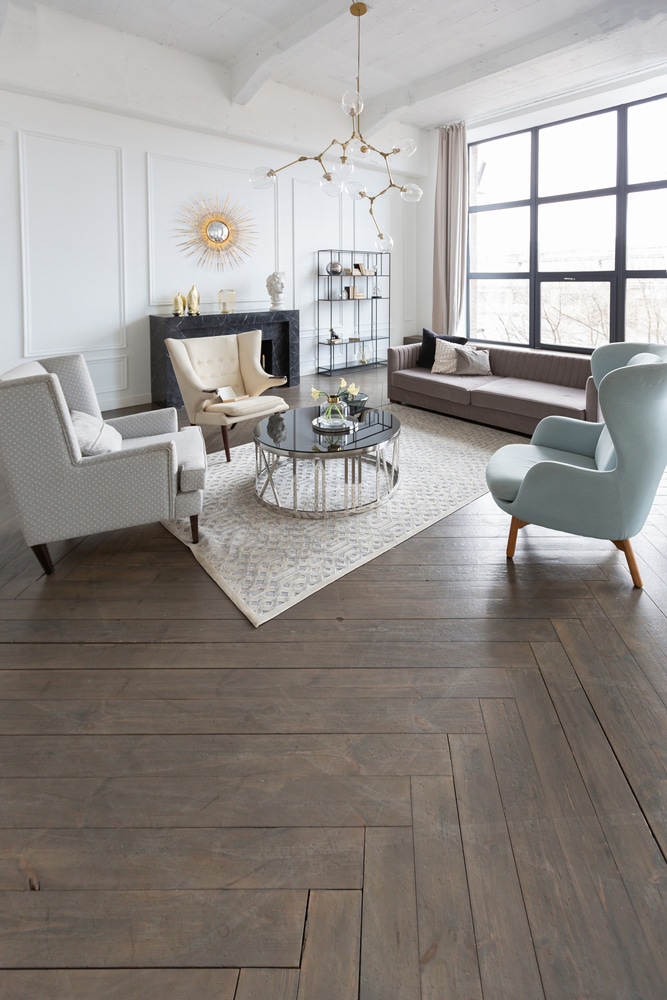 Salon design moderne luxe, parquet foncé et ambiance lumineuse. Salon moderne lumineux: murs blancs, parquet chevron gris foncé, cheminée marbre, chandelier design, fauteuils élégants et canapé taupe.