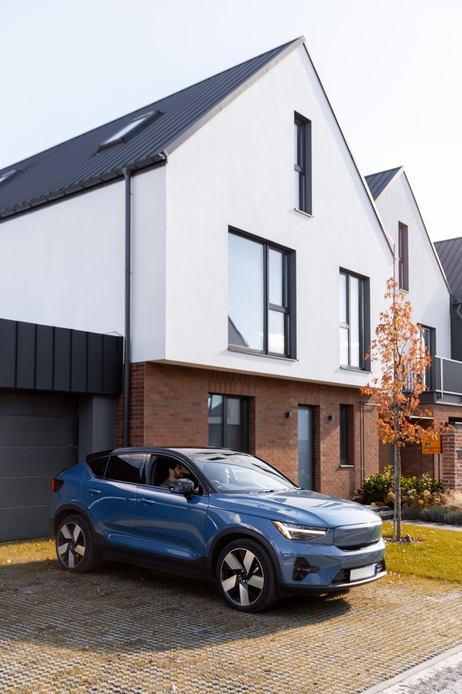 SUV électrique bleu devant une maison moderne design SUV électrique bleu garé devant une maison moderne blanche et brique, sous un toit noir. Allée en pavés drainants avec conducteur.