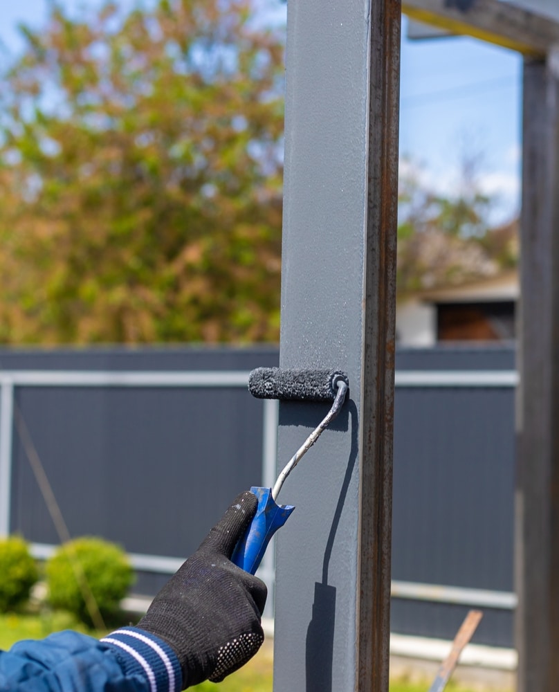 Peindre du métal au rouleau : conseils rénovation extérieure Main gantée appliquant de la peinture grise au rouleau sur un poteau métallique extérieur.