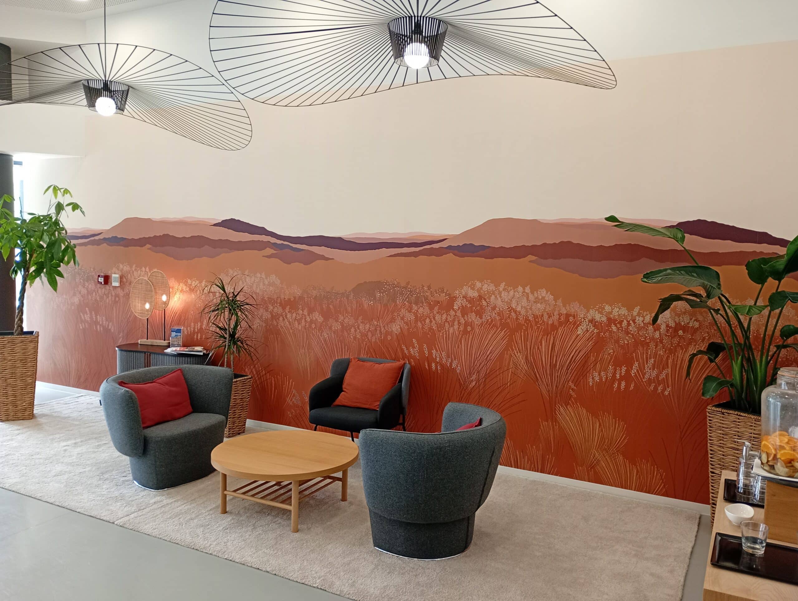 Décoration de salon moderne avec fresque murale désert Salon moderne avec murale désert orange, fauteuils gris et coussins rouges autour d'une table basse en bois.