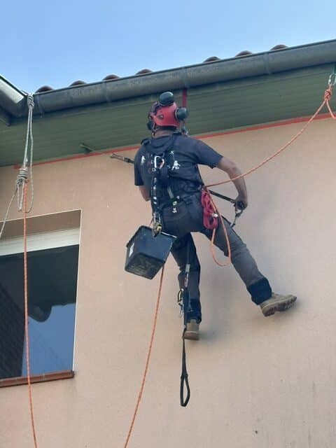 Cordiste professionnel effectuant des travaux de maintenance sur une façade de bâtiment avec équipement de sécurité.